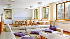 A modern living room with a sectional sofa, a wooden coffee table, and a dining area with tables and chairs. Large windows provide natural light and views of trees.