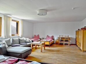 A modern living room with a sofa, a small table, a bed with pillows, a wooden cabinet, and a window with curtains.