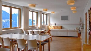 A spacious dining area with a long table and chairs, a sofa, and a kitchen area with a view of the mountains.