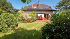 Una casa con techo inclinado, paneles solares, un patio con sillas rojas y un jardín bien cuidado.