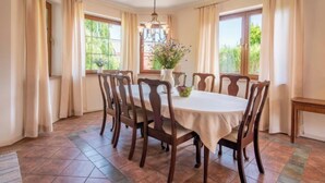 Comedor con una mesa redonda, seis sillas de madera, una lámpara de araña y ventanas grandes con cortinas.