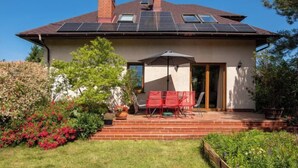Una casa con techo inclinado, paneles solares, una chimenea de ladrillo y un patio con sillas y mesa rojas.