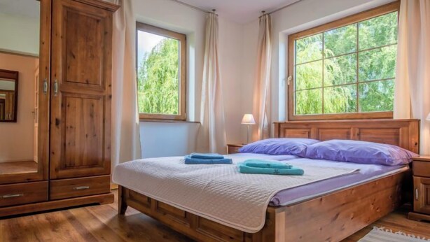 Una cama de madera con colcha blanca, dos toallas y un almohadón morado en una habitación con piso de madera, una ventana con vista a follaje y un armario de madera.