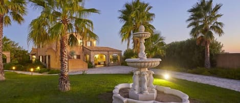 A landscaped garden with a fountain, palm trees, and a building with arched walkways.