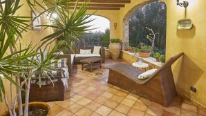 Un patio iluminado por el sol con muebles de mimbre, una planta grande en maceta y vistas de un jardín a través de un arco.