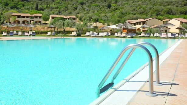 Una piscina con sillas de descanso y sombrillas, rodeada de edificios con montañas al fondo.
