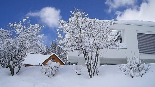 Une maison moderne avec un jardin enneigé, un chalet en bois et un ciel bleu dégagé.
