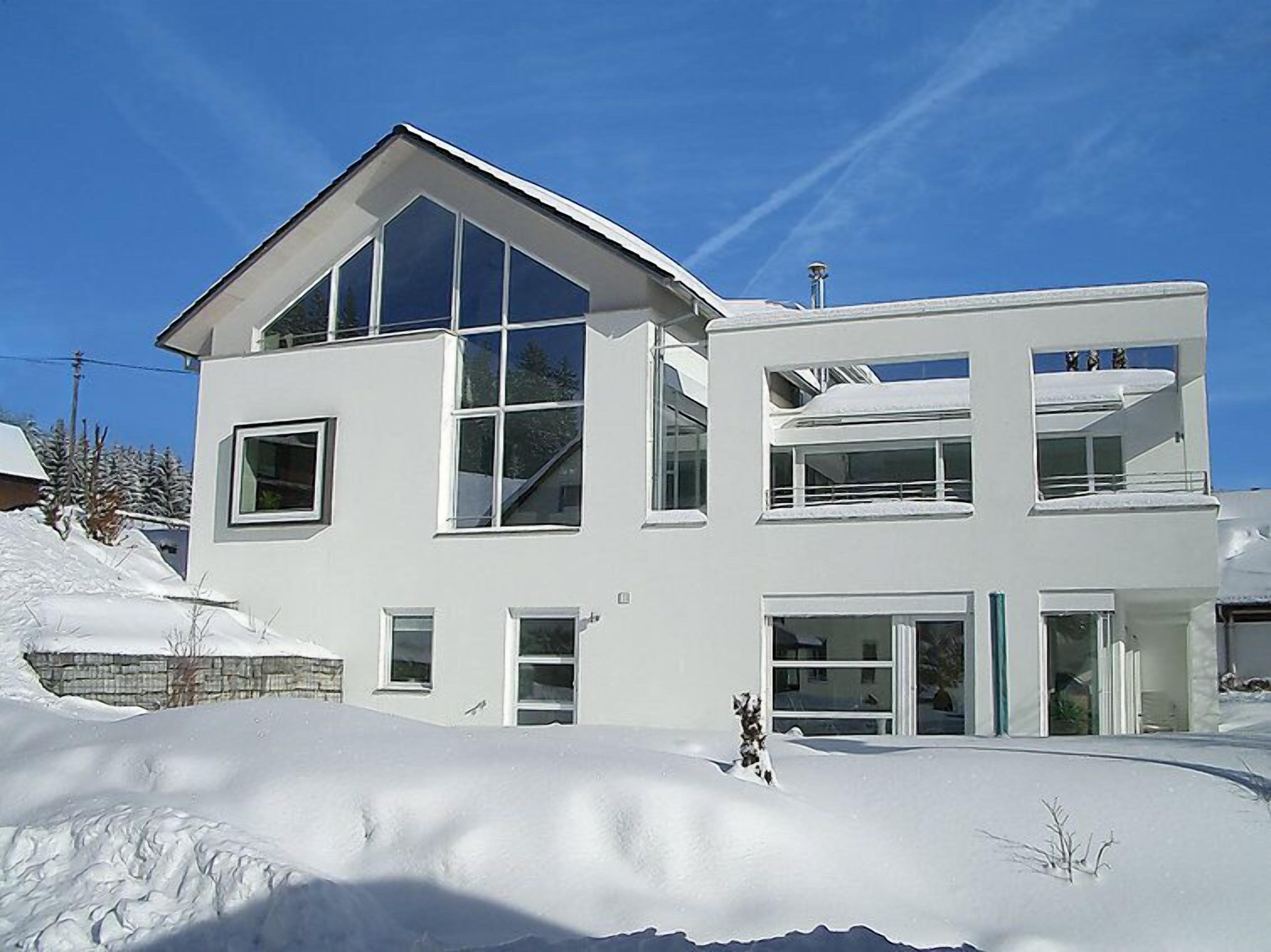Une maison moderne à deux étages, dotée de grandes fenêtres et d’un toit en pente, entourée de neige.