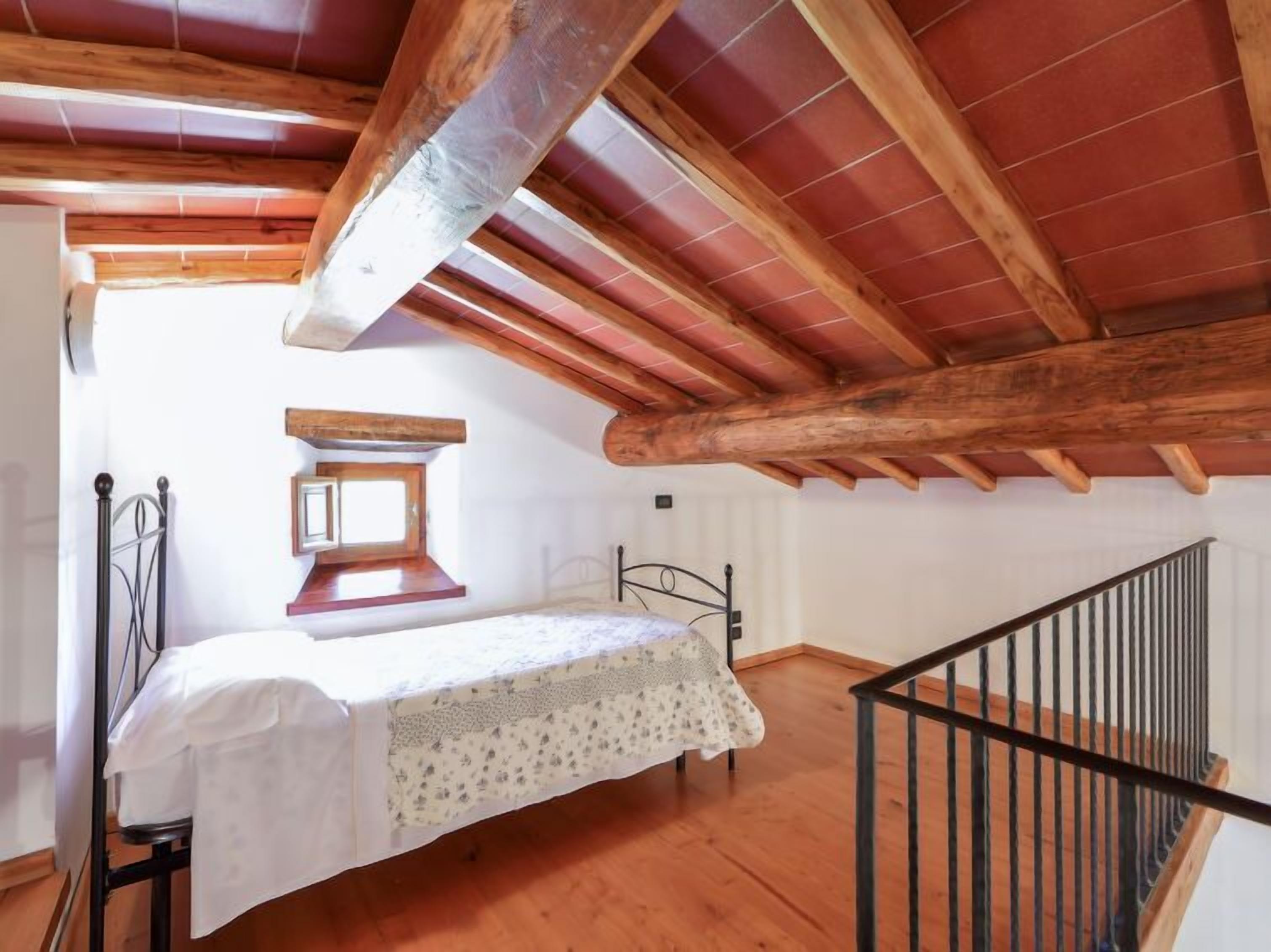 A bedroom with a wooden ceiling, a bed with a white bedspread, a small window, and a staircase.