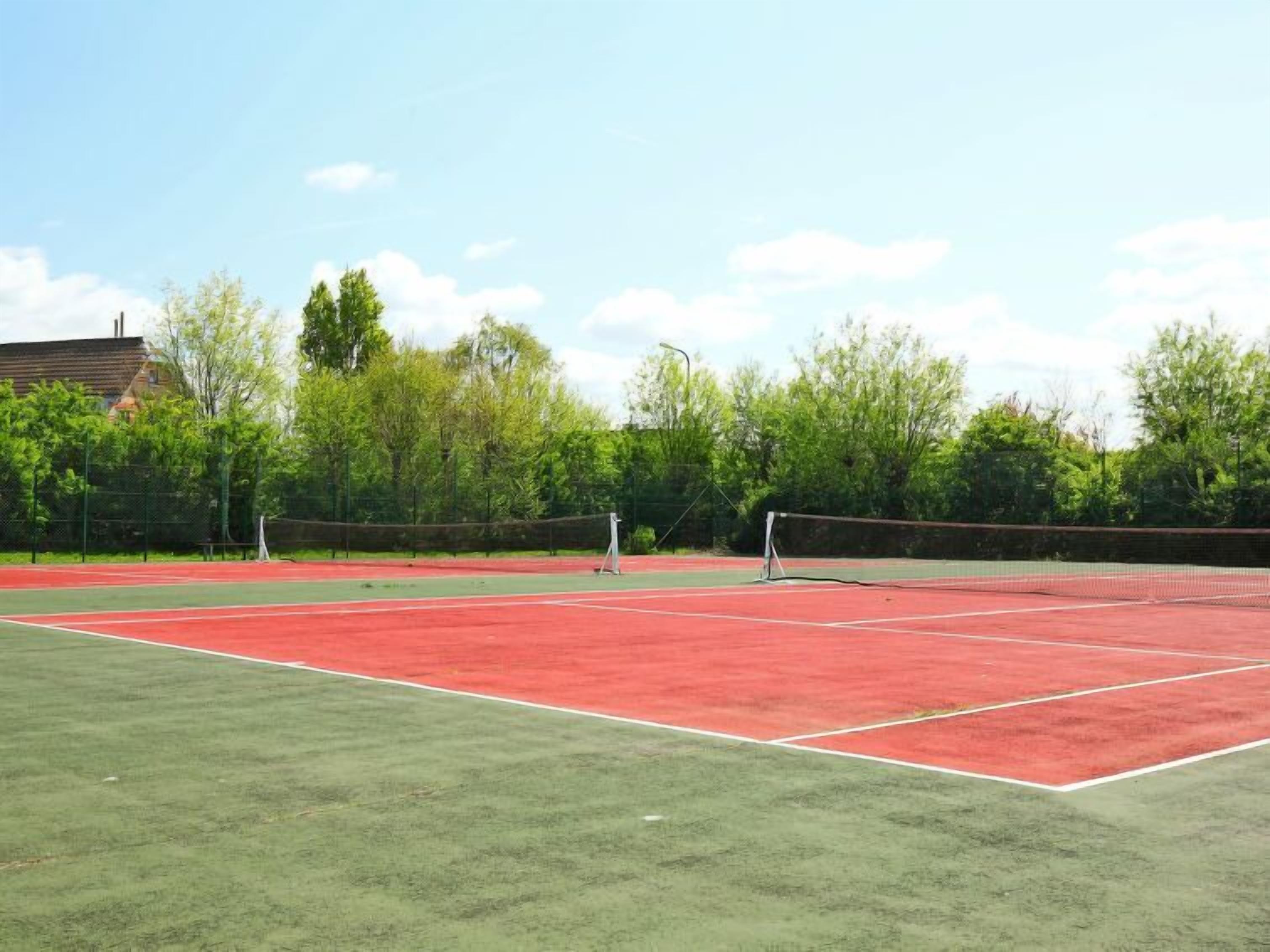 An outdoor tennis court with a net, surrounded by trees and a building in the background.