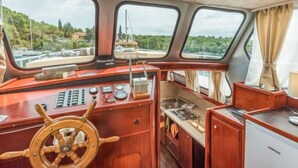 Un intérieur de bateau compact avec une console de commande en bois, une petite cuisine équipée d’une cuisinière et une vue sur une marina à travers trois fenêtres.