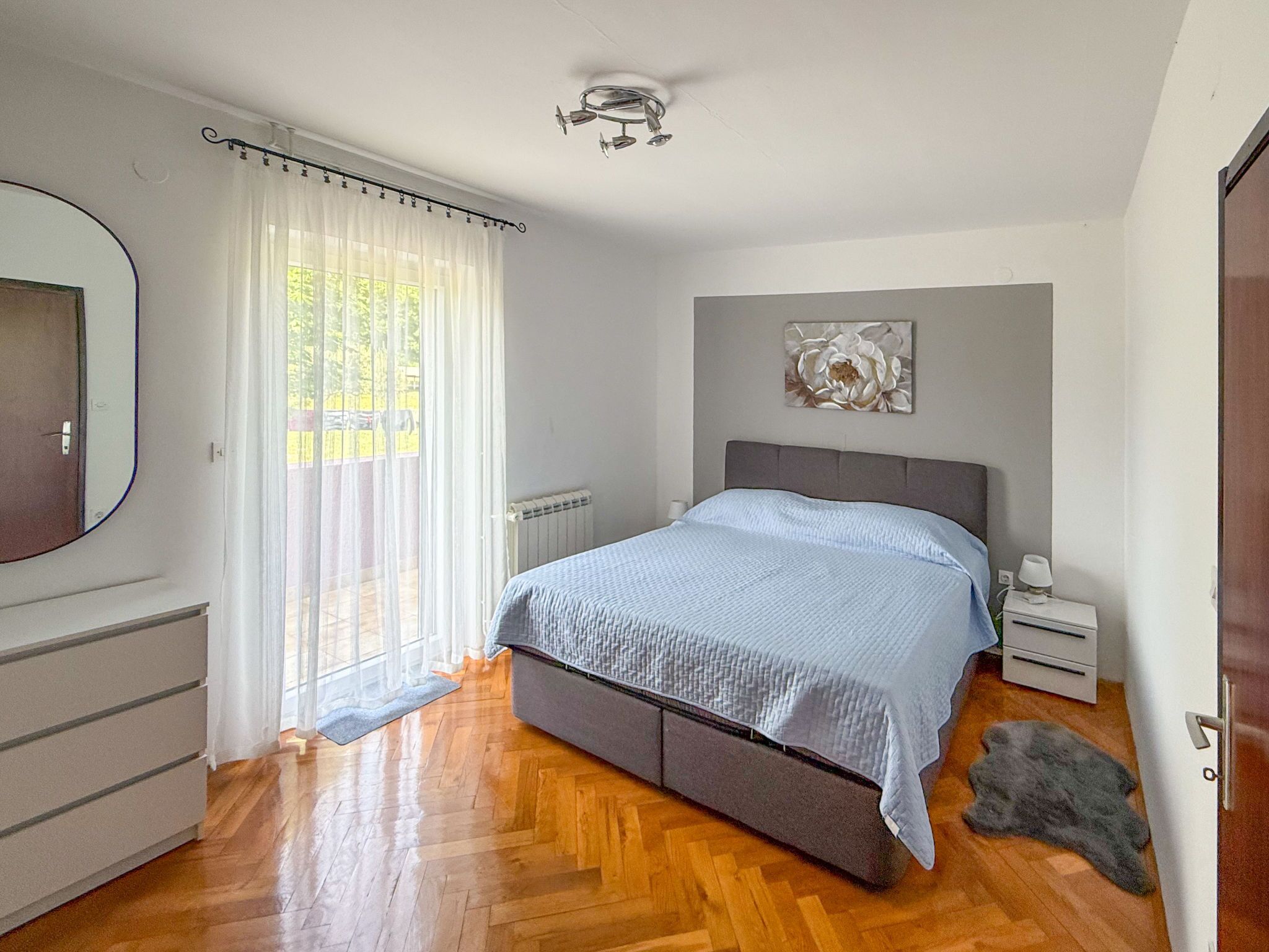 A bedroom with a bed, a bedside table, a nightstand, a radiator, a mirror, and a window with curtains.