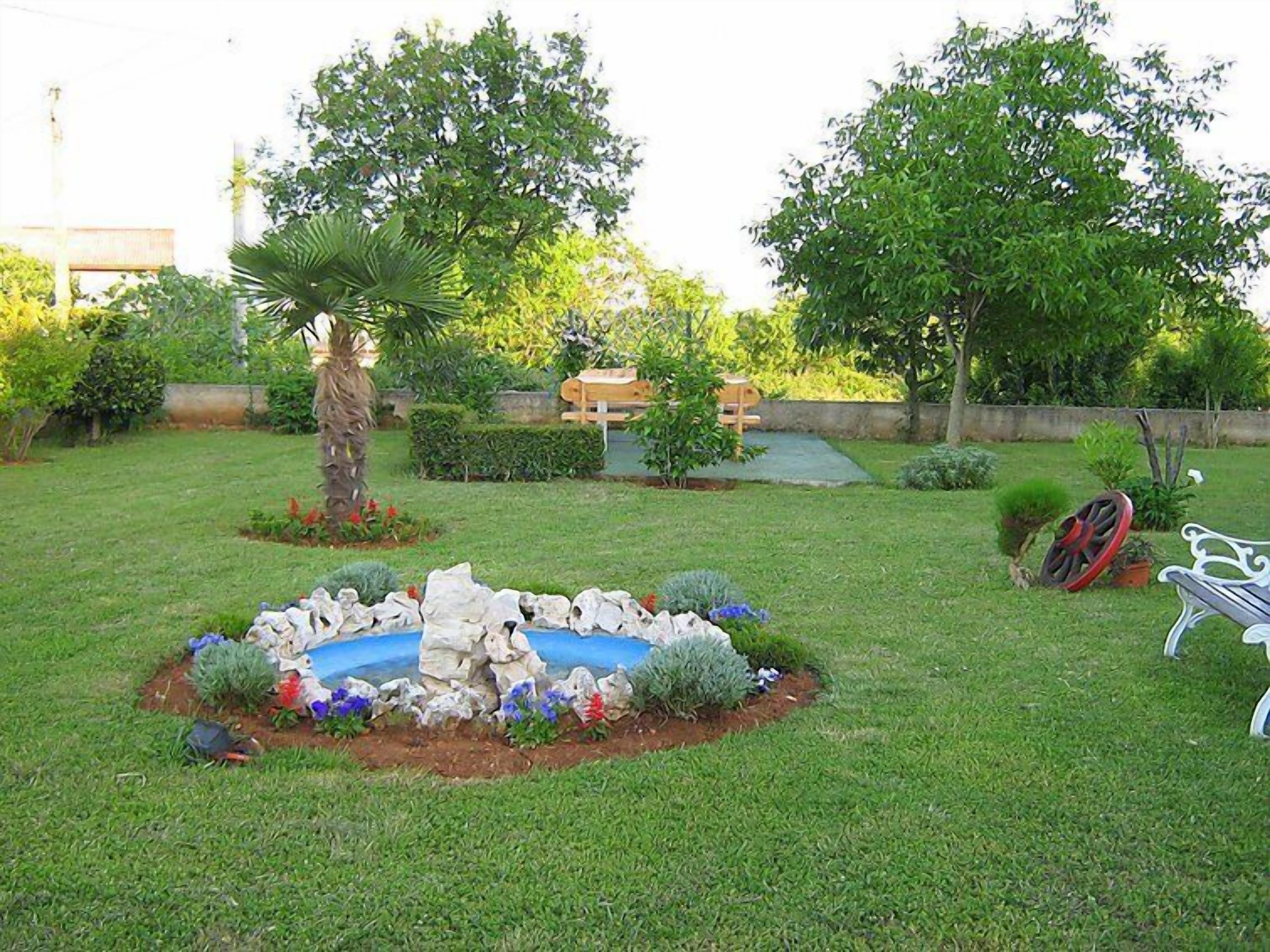 A well-maintained garden with a central rock feature surrounded by colorful flowers, a palm tree, and a bench.