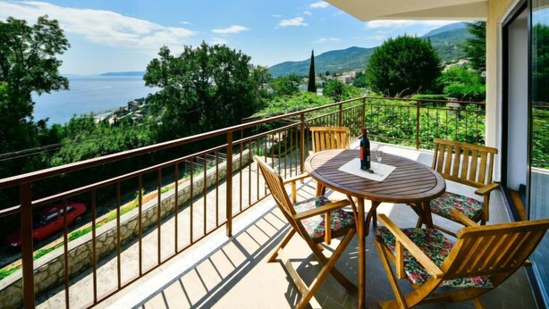 A balcony with a table and chairs, overlooking a scenic view of the sea and mountains.
