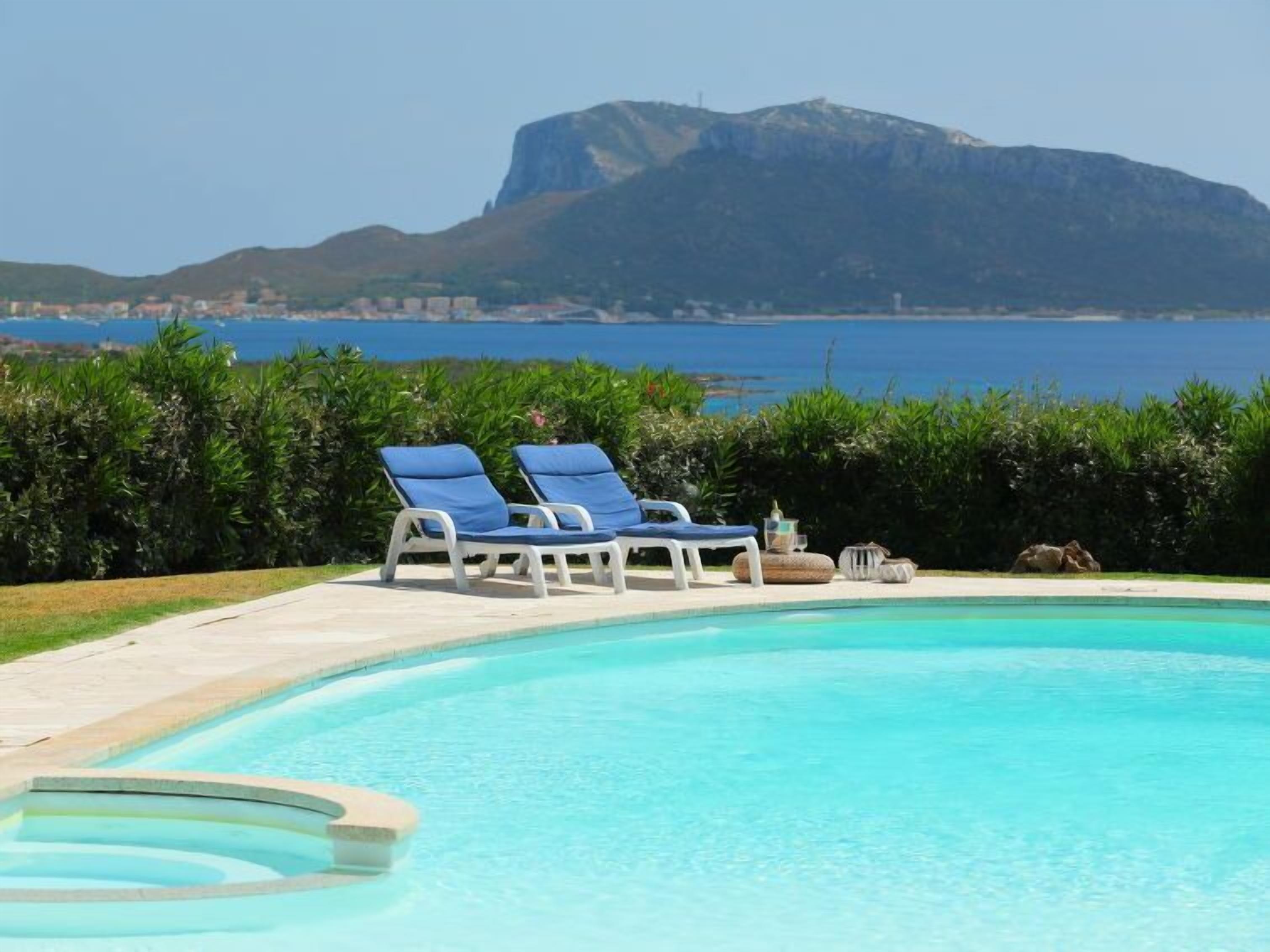 A swimming pool with two blue lounge chairs, a stone bench, and a view of a mountain and the sea.