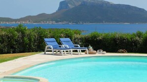 Piscina com duas espreguiçadeiras azuis, um banco de pedra e vista para uma montanha e o mar.