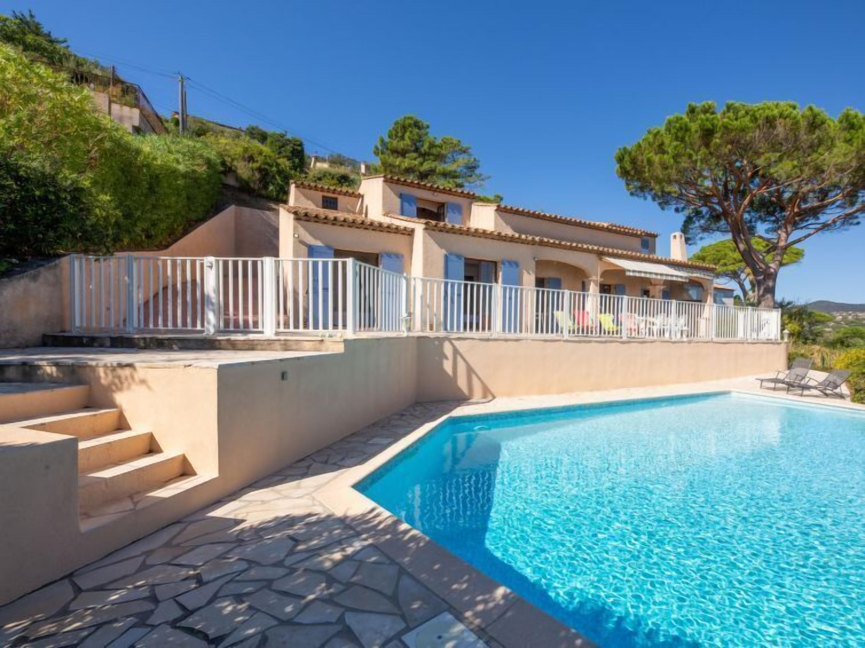 Blick auf einen Pool vor einer Villa im mediterranen Stil, mit einer Terrasse aus Steinplatten, einem Schwimmbecken und einem weißen Geländer.