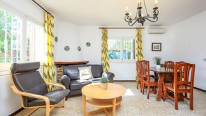 Ein helles Zimmer mit einer Couch, einem Esstisch mit Stühlen, einer Chandelier und einem Fenster mit Vorhängen.