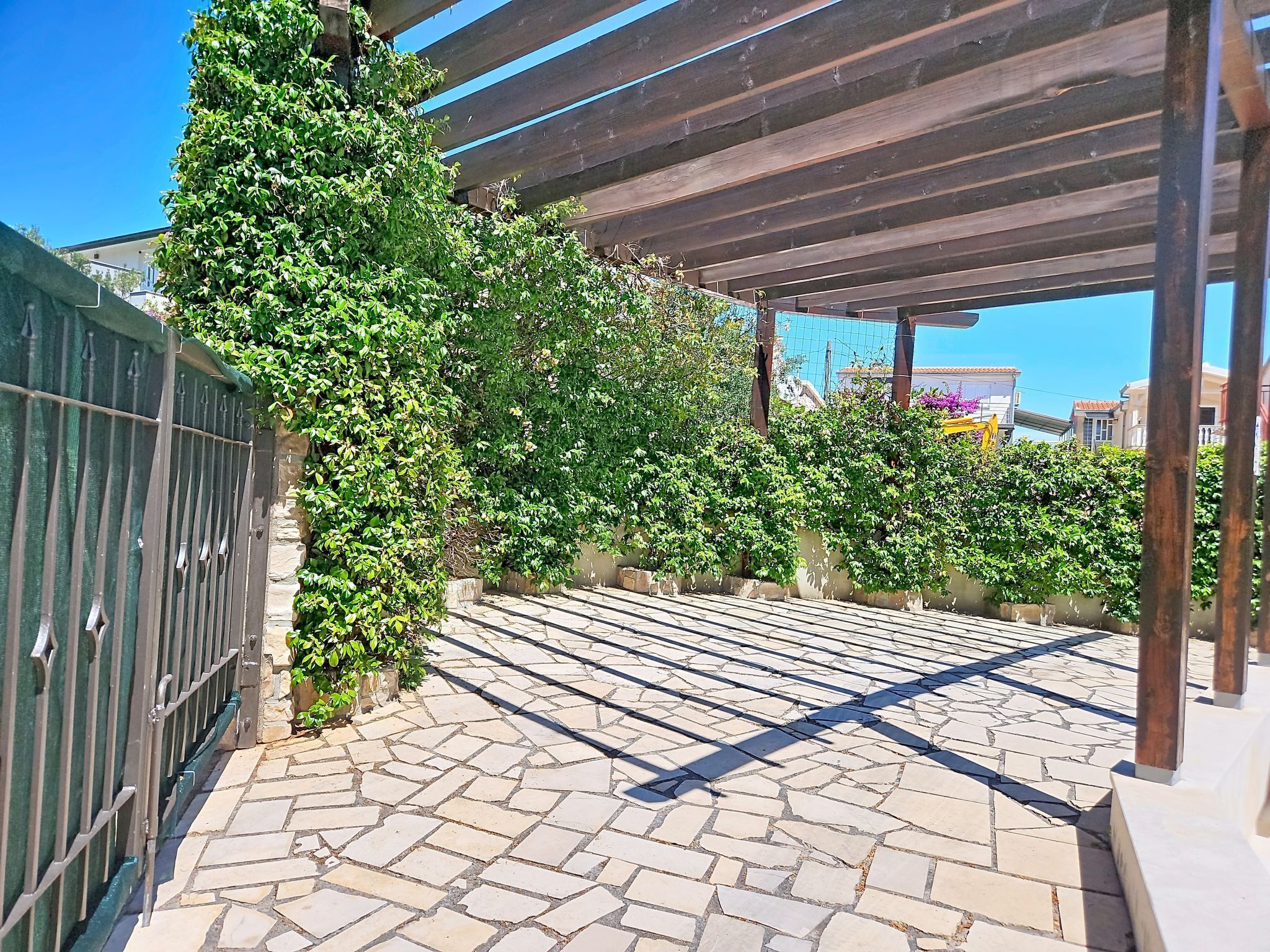 Ein gepflasterter Weg mit einer Pergola darüber, umgeben von üppigem Grün und einem Metallzaun.