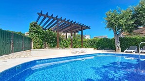 Une piscine avec un bord carrelé, une structure en forme de pergola et un espace clos agrémenté de verdure.