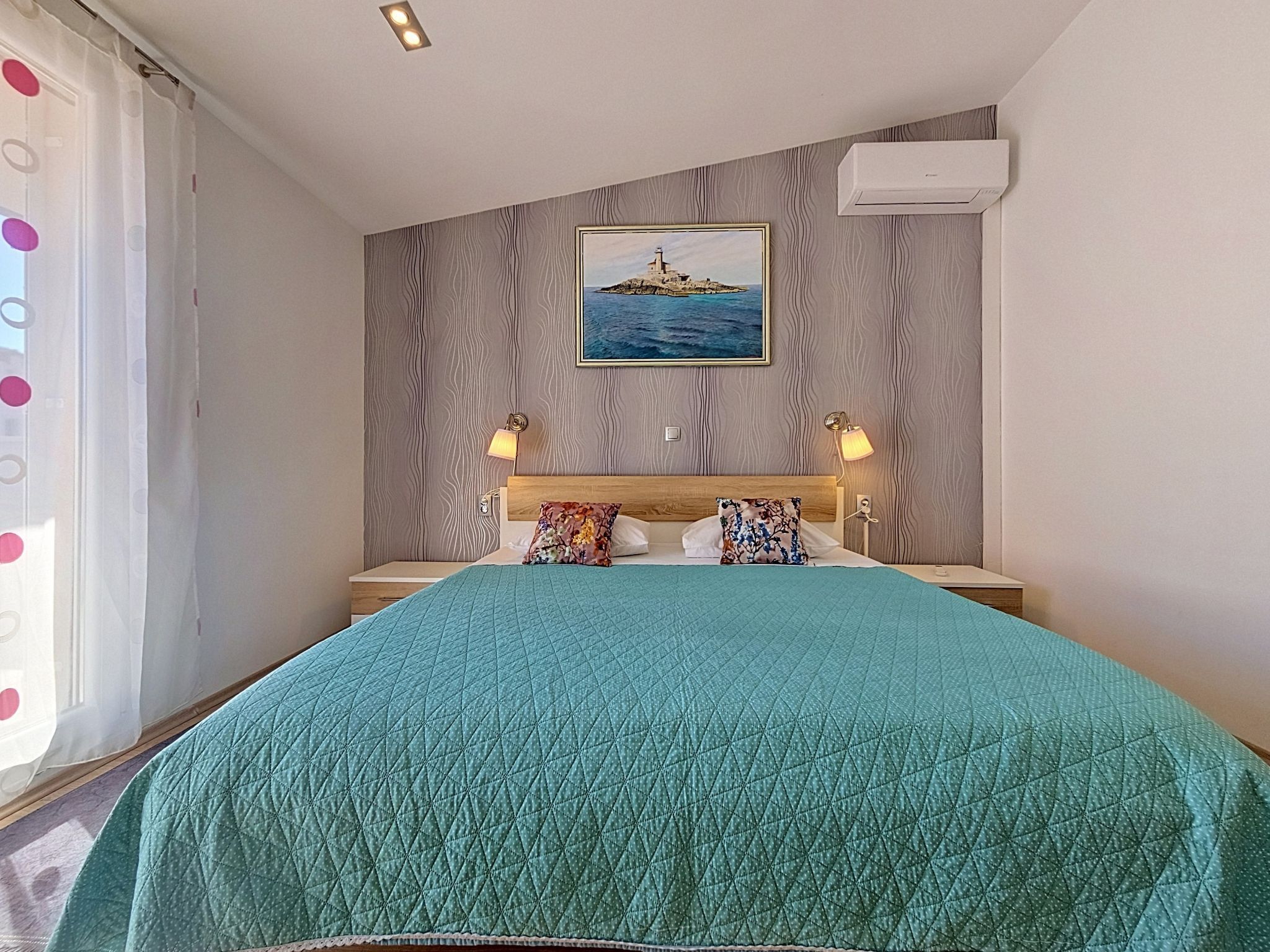 Une chambre à coucher comprenant un lit, une tête de lit, une table de chevet, une lampe, un tableau au mur et un appareil de climatisation.