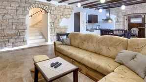 Sala de estar com parede de pedra, vigas de madeira, um sofá, uma mesa com controles remotos e uma cozinha ao fundo.