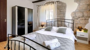 Quarto com cama, guarda-roupa, parede de pedra e janela com cortinas.