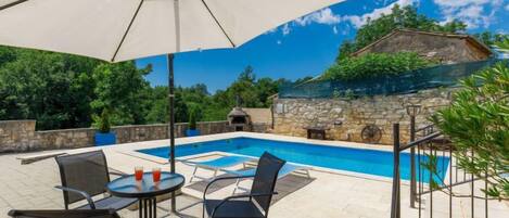 A pool area with a table and chairs, a large umbrella, and a stone wall in the background.