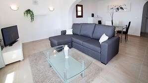 Een moderne woonkamer met een grijze hoekbank, een glazen salontafel, een flatscreen-tv en een eethoek met een tafel en stoelen.