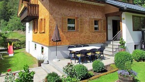 Ein zweistöckiges Holzhaus mit Balkon, einer Terrasse mit Tisch und Stühlen sowie einem Garten mit verschiedenen Pflanzen.