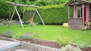 Ein Holzschaukel, ein kleines Holzspielhaus und ein gepflegter Garten mit verschiedenen Pflanzen und einem Hecken im Hintergrund.