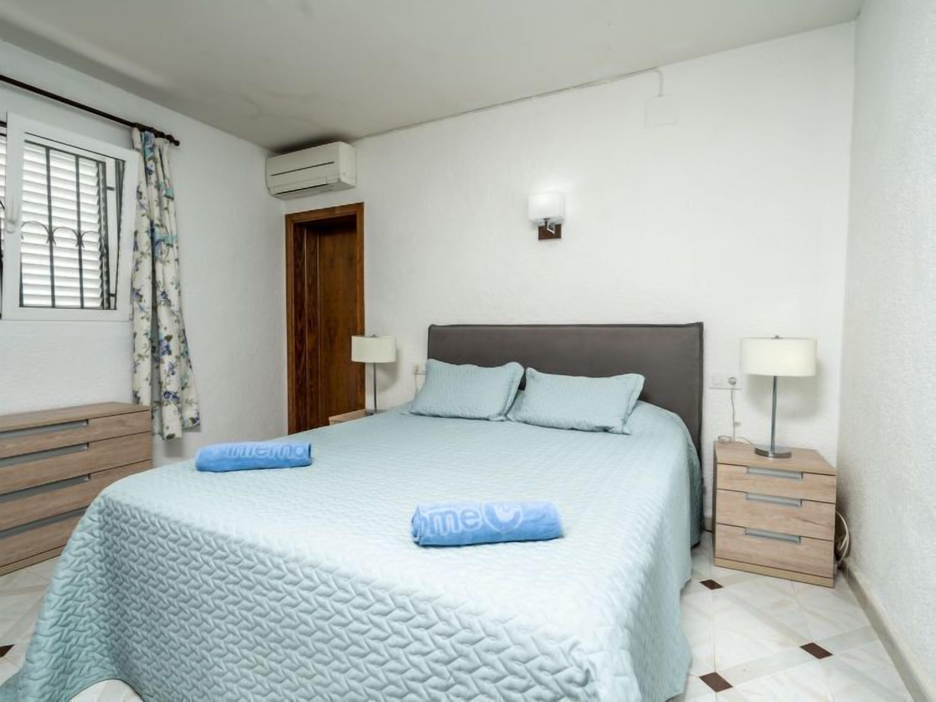 Une chambre à coucher comprenant un lit, deux tables de chevet, une commode, un appareil de climatisation, une fenêtre avec des rideaux et un luminaire fixé au mur.
