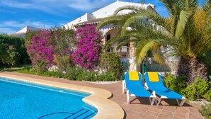 Un espace aménagé au bord d’une piscine à l’eau d’un bleu limpide, avec des chaises longues et un bâtiment blanc doté de balcons.