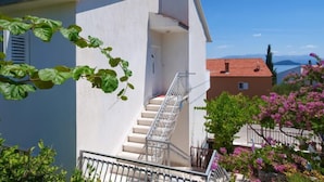 Un bâtiment blanc avec un escalier menant à un balcon, entouré de verdure et surplombant un ciel bleu clair.