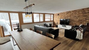 A spacious living area with a wooden dining table, sofas, and a TV. Large windows provide natural light and a view of the outdoors.