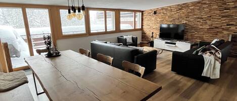 Een ruime woonkamer met een houten eettafel, banken en een televisie. Grote ramen zorgen voor natuurlijk licht en uitzicht op de omgeving.