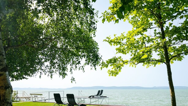 Blick auf einen See mit zwei Liegestühlen und einem Picknicktisch auf einer Wiese.