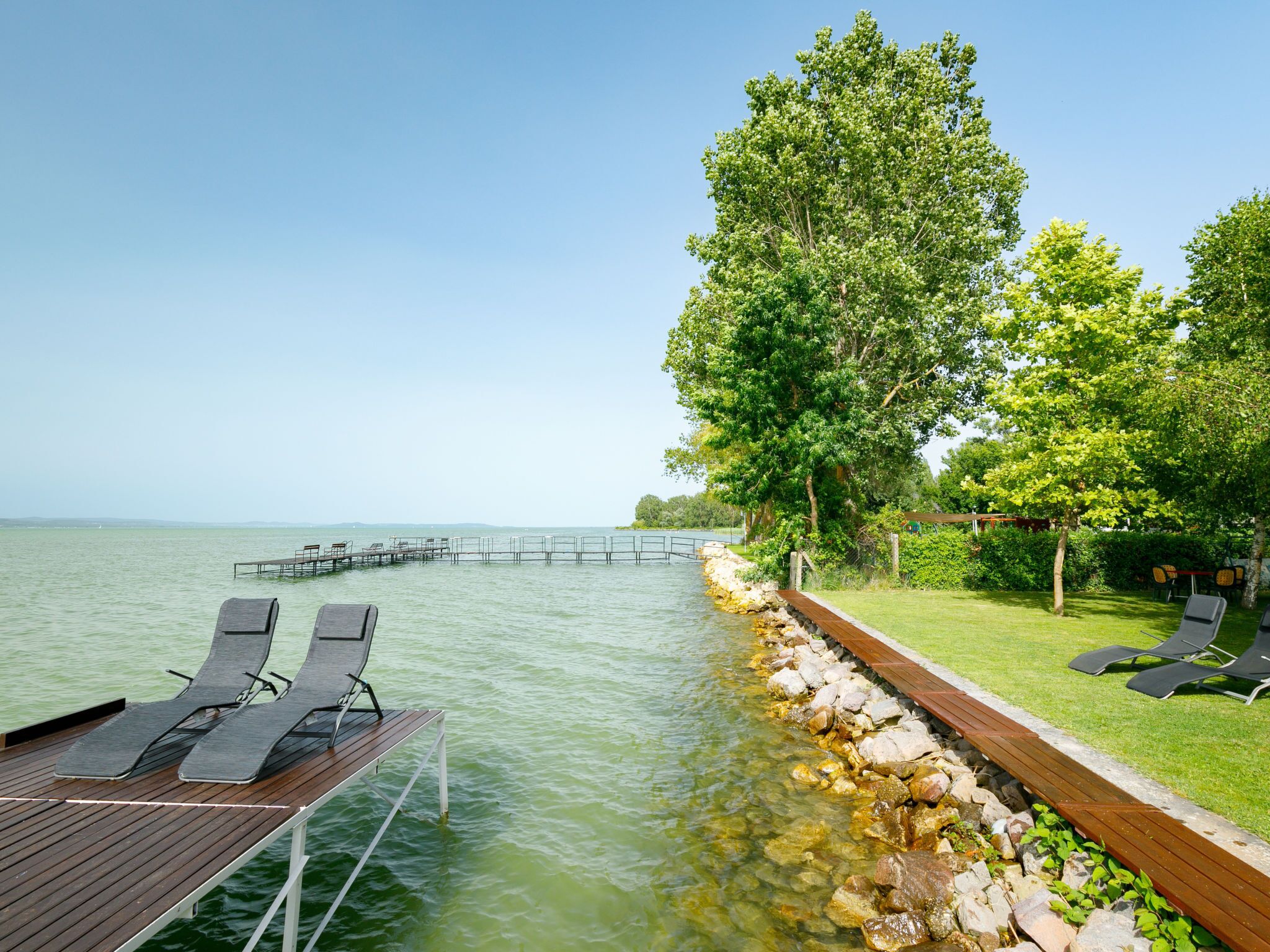 Een houten terras met lounge stoelen loopt uit over een rotsachtige kustlijn in kalm water, met een gazon, bomen en een heldere lucht.