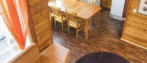 A wooden dining area with a table and chairs, a small kitchen with a refrigerator, and a window with curtains.