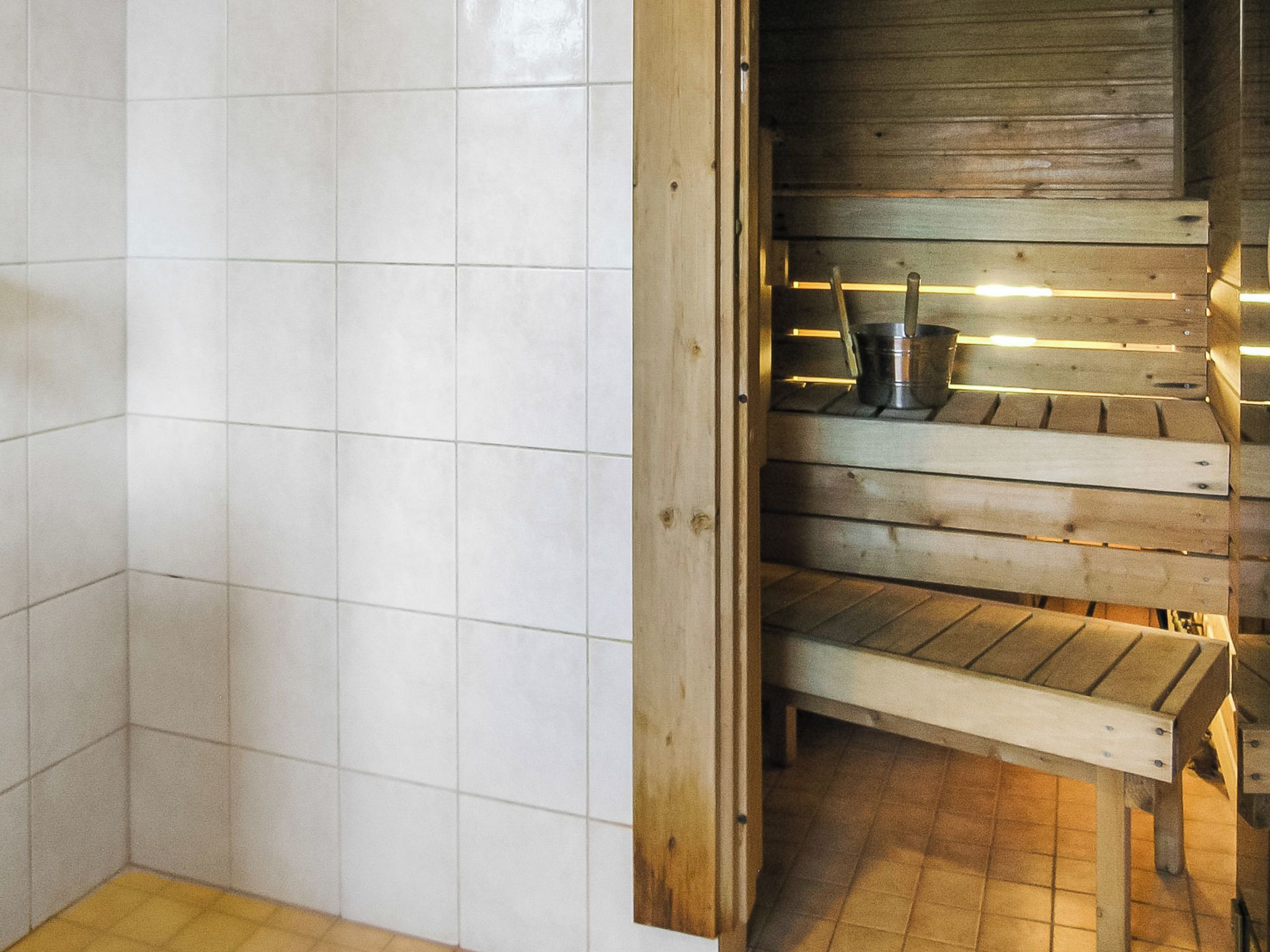 A sauna with wooden benches and a metal bucket, surrounded by white tiled walls.