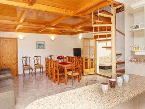 A cozy room with a wooden ceiling, a dining area with wooden chairs and a table, a sofa, a TV, and a staircase leading to another level.