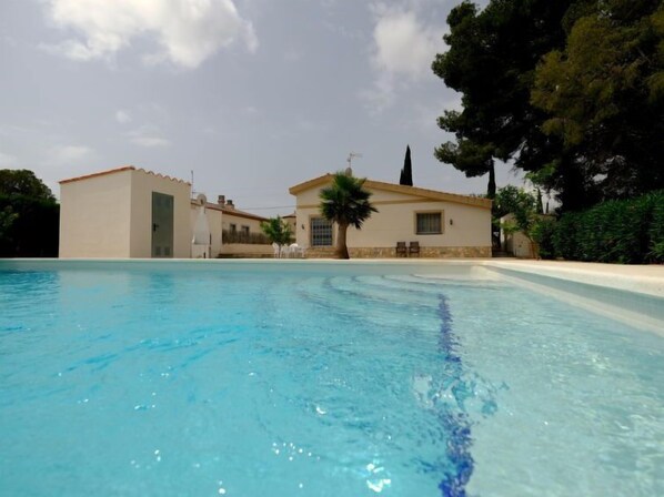 A swimming pool with a clear blue water surface, surrounded by a building with a tiled roof and a palm tree.