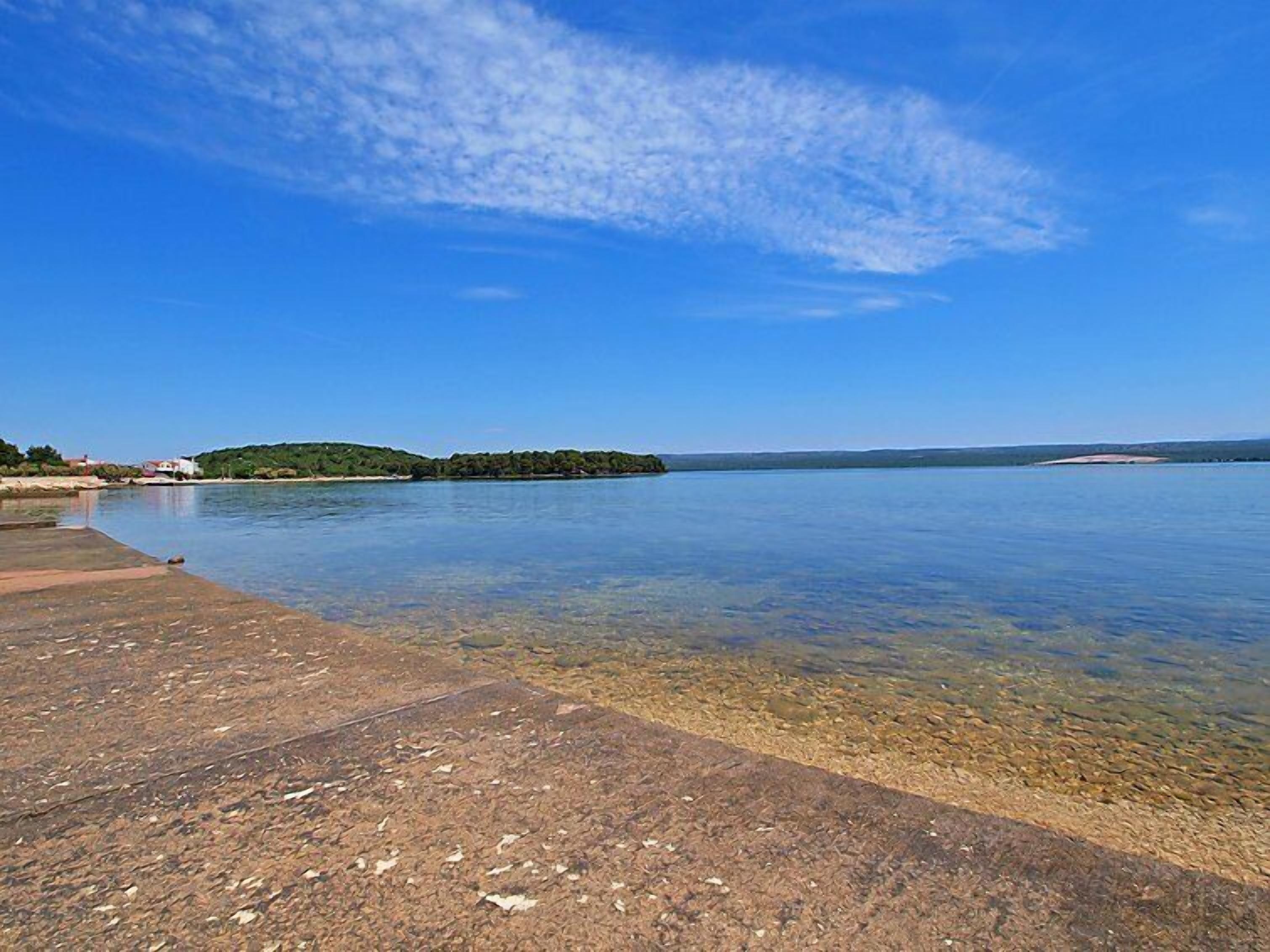 On peut admirer un lac paisible, avec une petite île qui se dessine à l’horizon, un quai en béton et un ciel bleu dégagé.