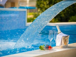A poolside setting with a water fountain, champagne bottle, glasses, and fresh fruits.