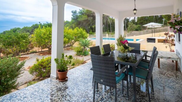 Un patio cubierto con una mesa y sillas, una piscina al fondo y vistas del paisaje circundante.