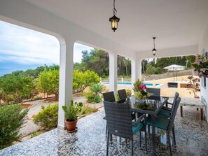 Une terrasse couverte avec une table et des chaises, une piscine à l’arrière-plan et une vue sur le paysage environnant.