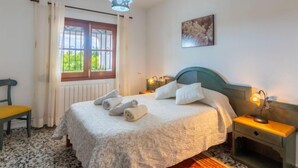 Un dormitorio con cama, ventana con cortinas, mesita de noche con lámpara y una silla.