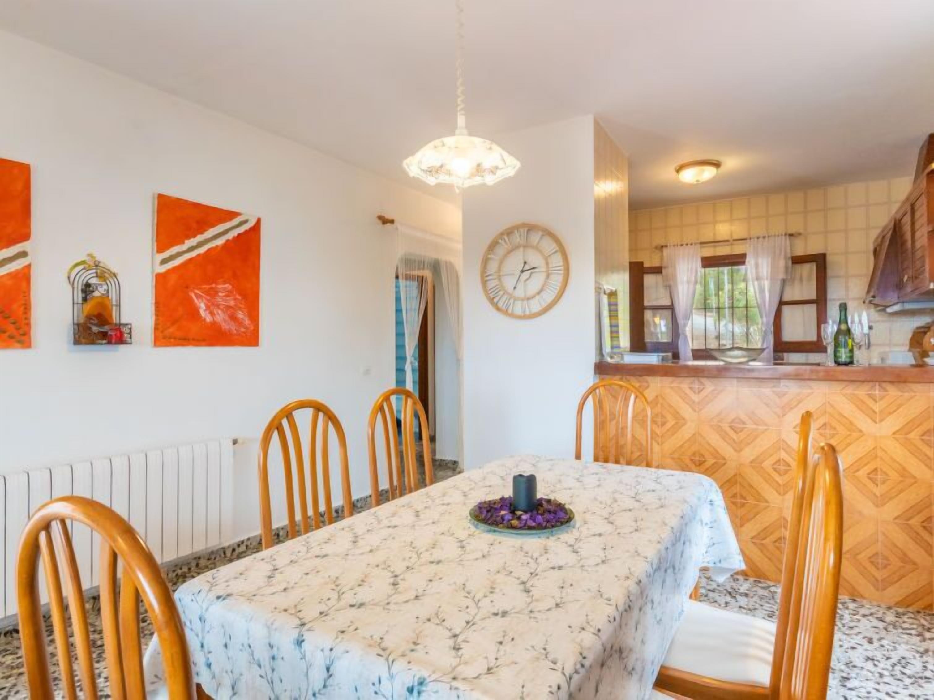 Un comedor con una mesa preparada para cuatro personas, sillas de madera, un reloj en la pared y una cocina visible al fondo.