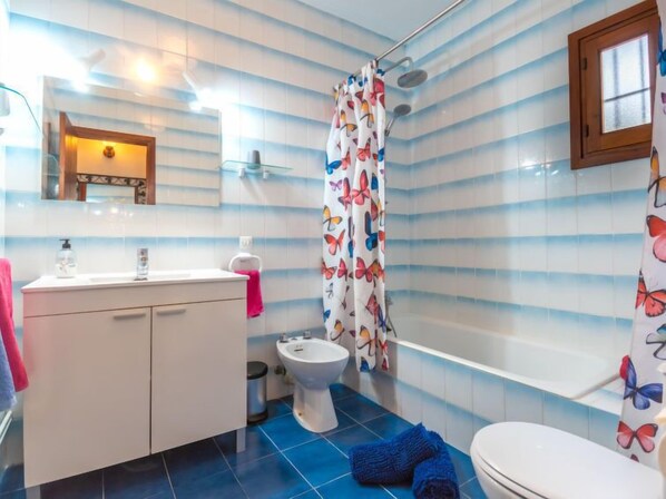 Ein Badezimmer mit einer Wand aus gestreiften Fliesen, einem weißen Waschbecken mit Spiegel darüber, einer Toilette, einem Bidet und einem Duschvorhang mit Schmetterlingsmuster.