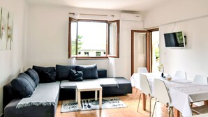 Un salon moderne comprenant un canapé, une petite table, un coin repas avec des chaises, un téléviseur fixé au mur et une fenêtre avec un volet ouvert.
